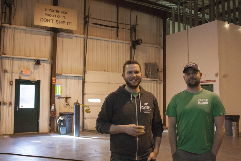 Castle Island Brewing Company founder and owner Adam Romanow (left) and head brewer Matt DeLuca stand in the brewery, below a sign that reads, "If you're not proud of it ... don't ship it!" The sign was in the building before Romanow signed the lease for the space and convinced him that this was the right spot to start Castle Island Brewing Company. (Photo by Susan Doucet)